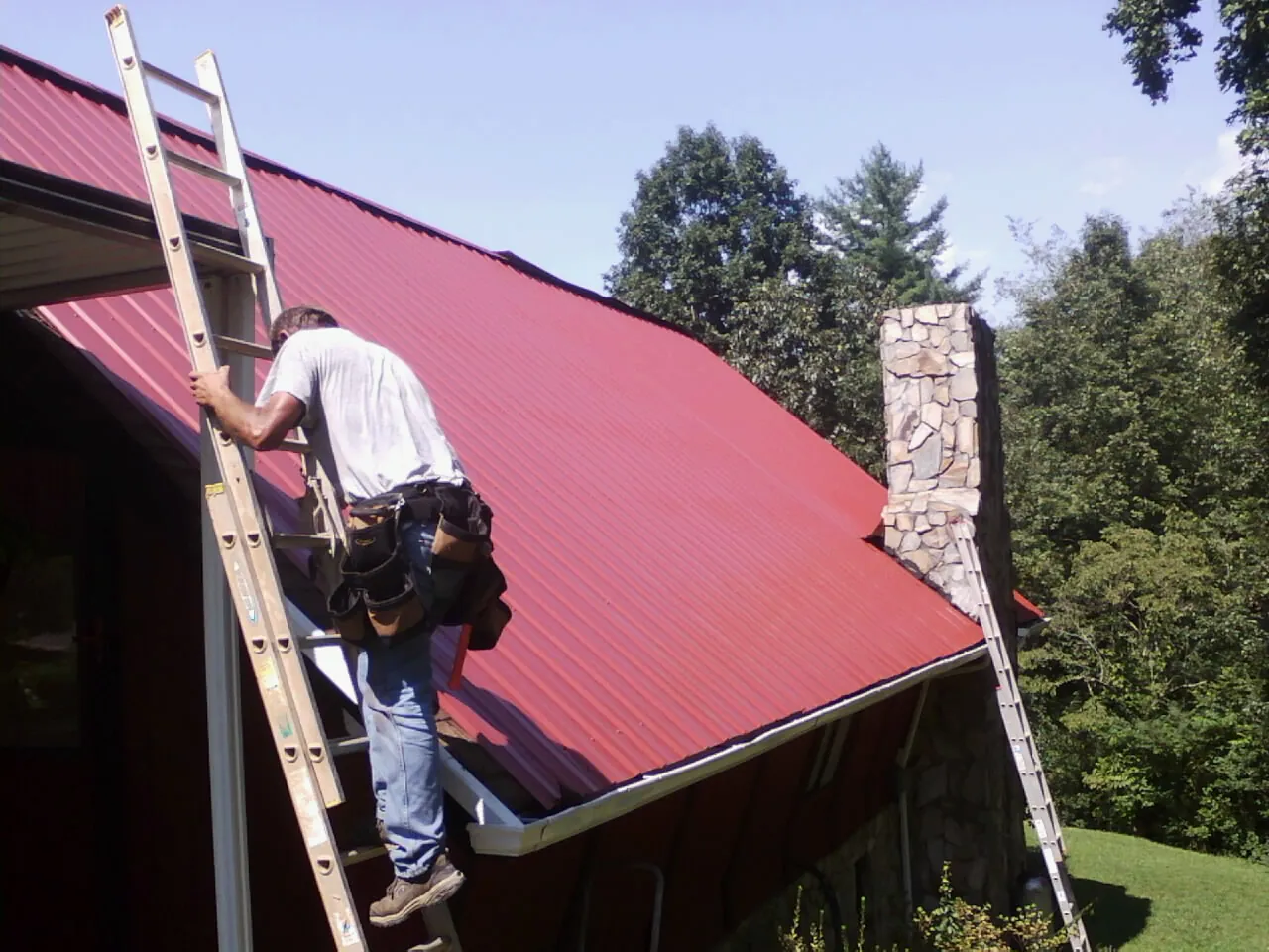 Roofer on a ladder beside a red metal roof and stone chimney — Soffit & Fascia Repair by Roofing Brothers in Lehigh